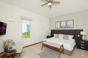 Une chambre à coucher moderne, avec un lit, des tables de chevet, un ventilateur de plafond, une fenêtre et des œuvres d’art accrochées au mur.