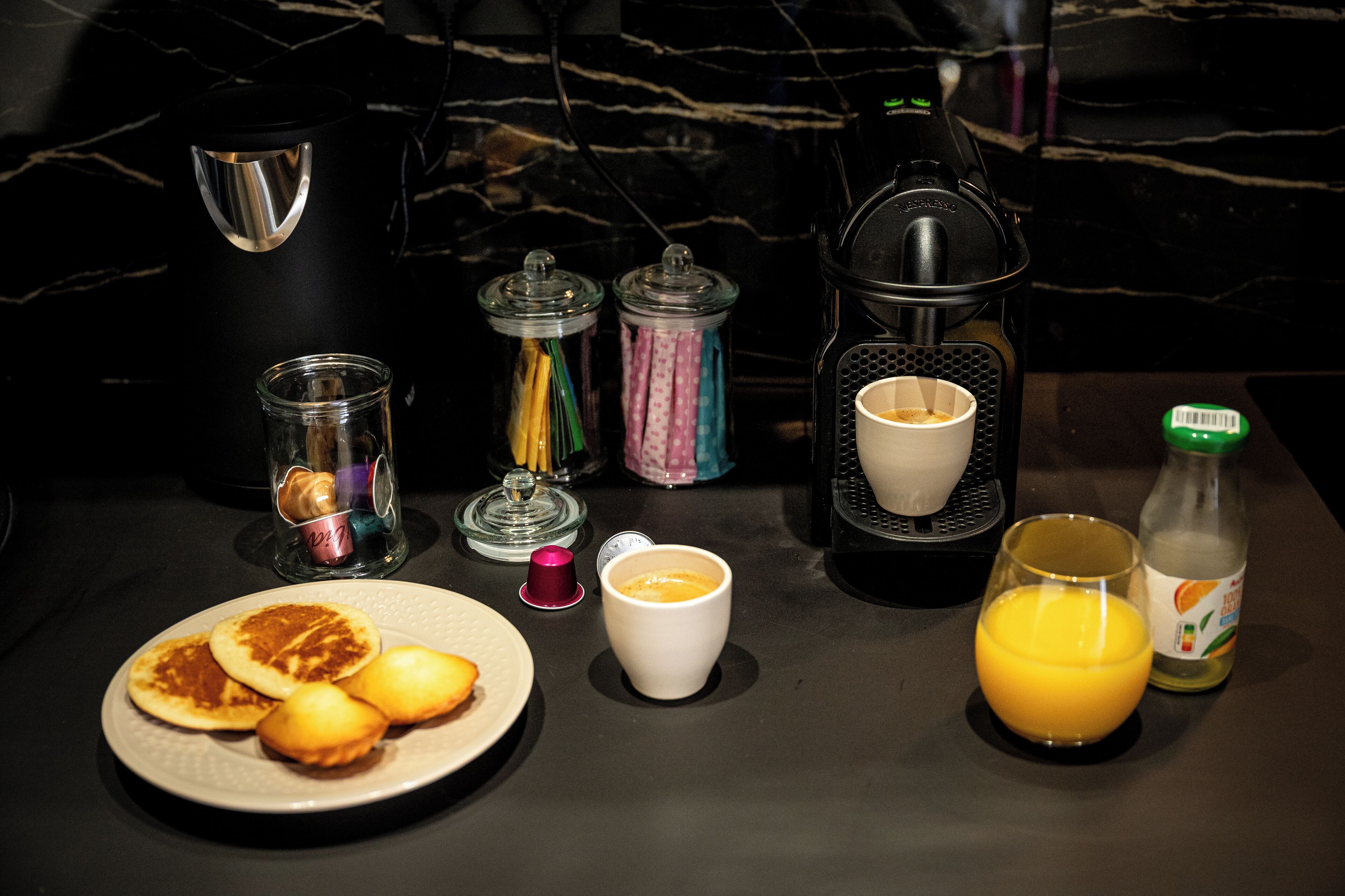 A breakfast setting with pancakes, a coffee maker, a glass of orange juice, and various condiments.