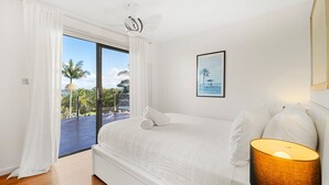 A bedroom with a bed, a ceiling fan, a framed picture, and a view of palm trees and a beach through a sliding glass door. - Lakeviews - Pet Friendly with Fireplace (Culburra Beach)