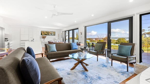 A modern living room with a sofa, armchairs, and a coffee table. There is a kitchen area with cabinets and a dining table. Large windows offer a view of the outdoors. - Lakeviews - Pet Friendly with Fireplace (Culburra Beach)