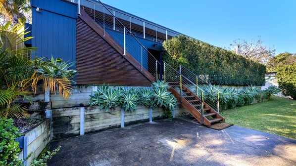 Une maison moderne avec un escalier en bois menant à un balcon, entourée d’une végétation luxuriante et d’une pelouse bien entretenue.