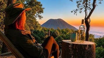 Uma pessoa sentada em uma cadeira ao ar livre, com vista para uma montanha e uma garrafa sobre um toco de árvore.