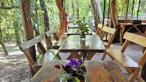 An outdoor wooden table with a glass vase of flowers, surrounded by benches and tables in a forested area.