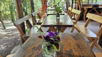 Mesa de madeira ao ar livre com um vaso de vidro cheio de flores, cercada por bancos e mesas em uma área arborizada.