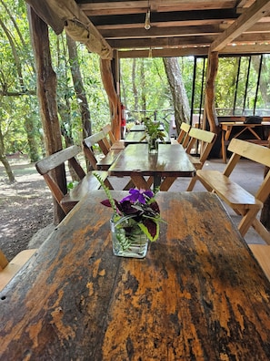 An outdoor wooden table with a glass vase of flowers, surrounded by benches and tables in a forested area.