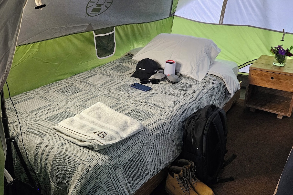 A tent with a bed, a pillow, a cup, a cap, a backpack, and a pair of boots.
