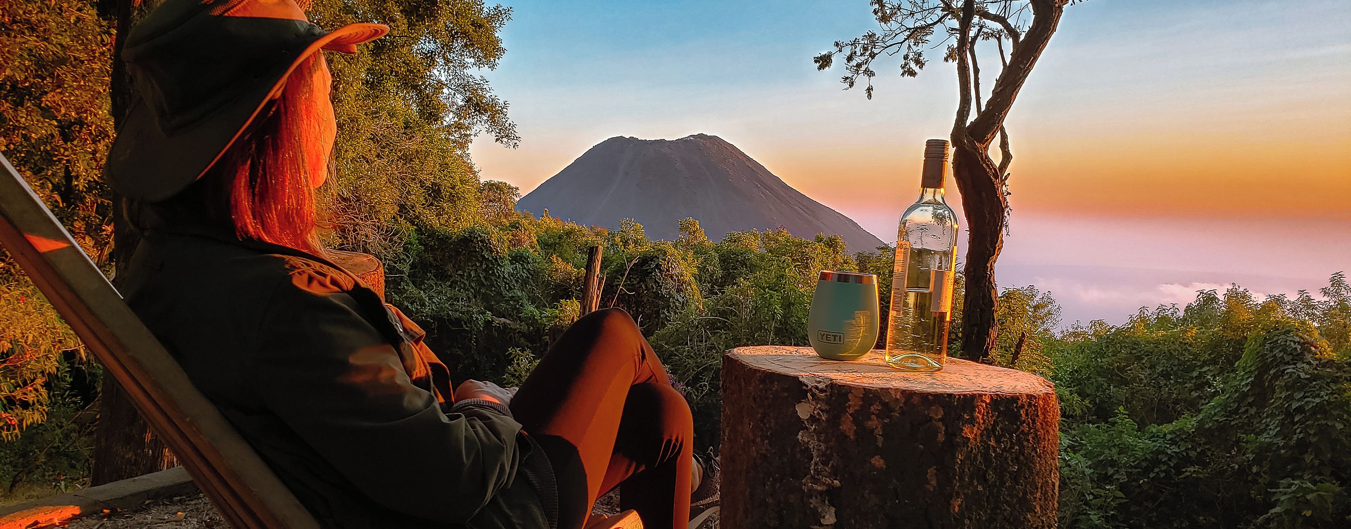 A person sitting on a bench overlooking a mountain, with a bottle and glass on a tree stump.
