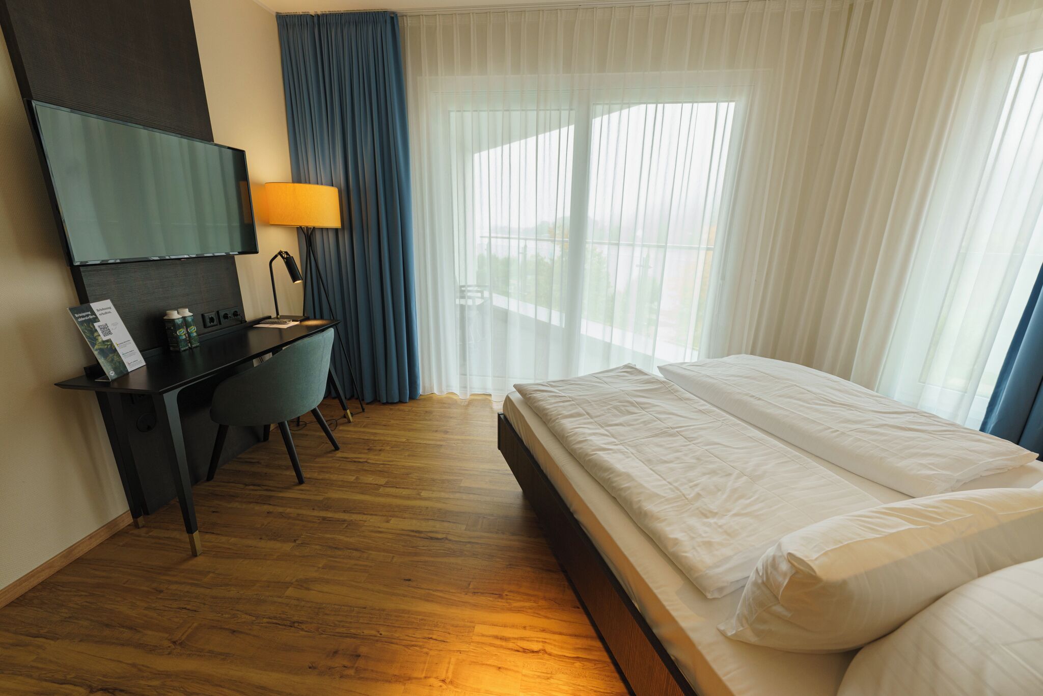 A hotel room with a bed, a desk with a chair, a TV, and a window with curtains.