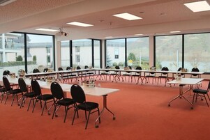 Sala de conferências com várias mesas e cadeiras, amplas janelas e piso coberto de carpete.