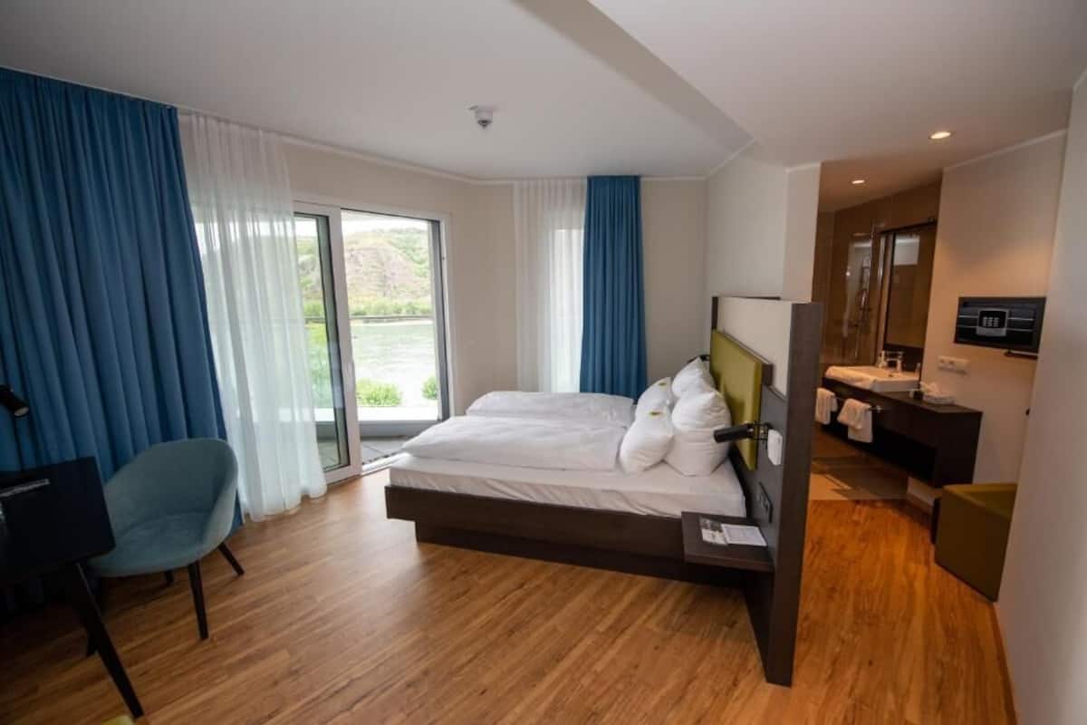 Ein modernes Hotelzimmer mit einem großen Bett, einem Schreibtisch, einem Stuhl, einem Badezimmer mit Spiegel und Waschbecken sowie Blick ins Freie.