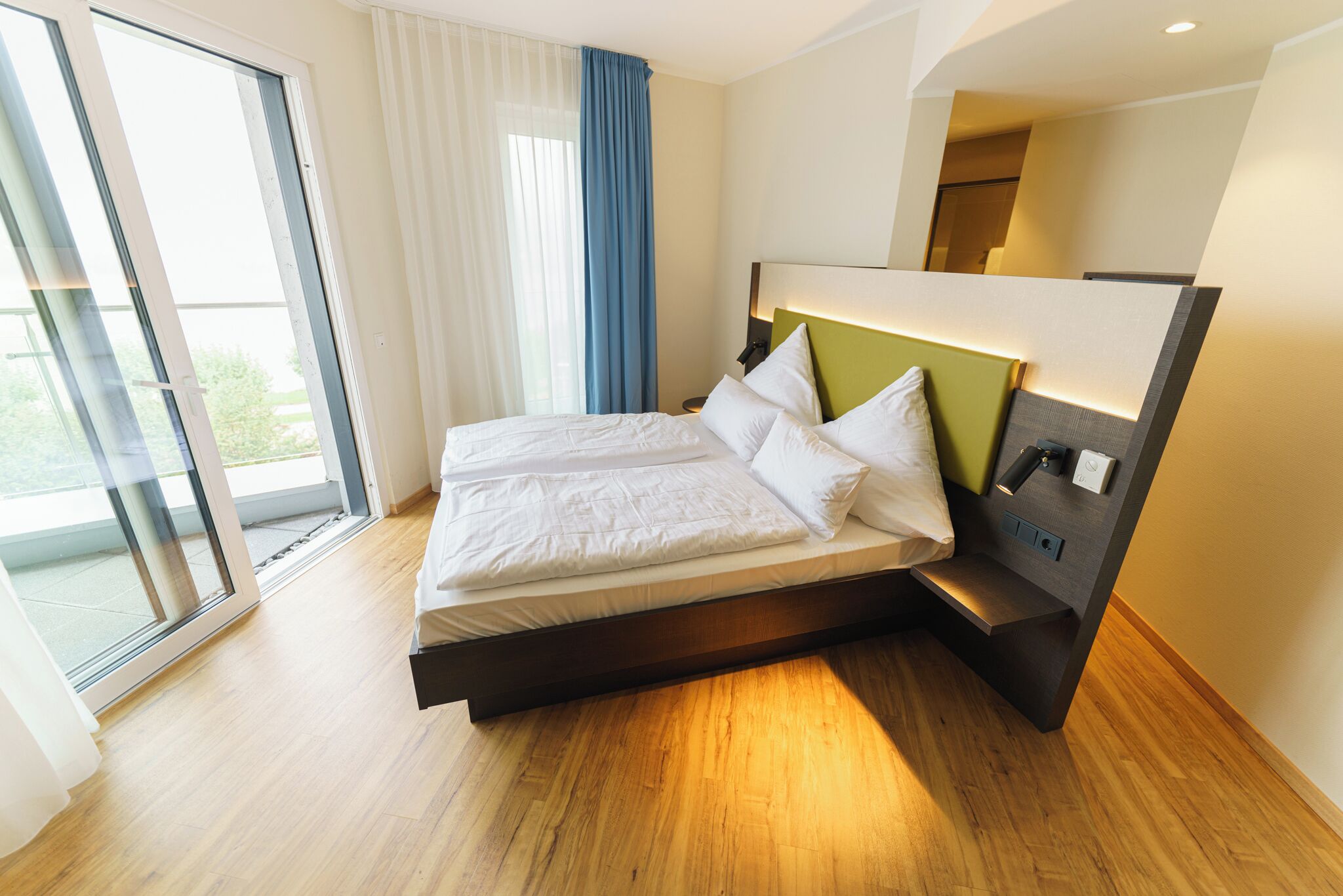 A modern hotel room with a large bed, a bedside table, and a view through a sliding glass door.