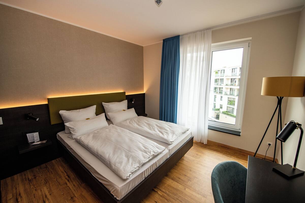 Ein Hotelzimmer mit Bett, Schreibtisch, Lampe und einem fenster mit Vorhängen.