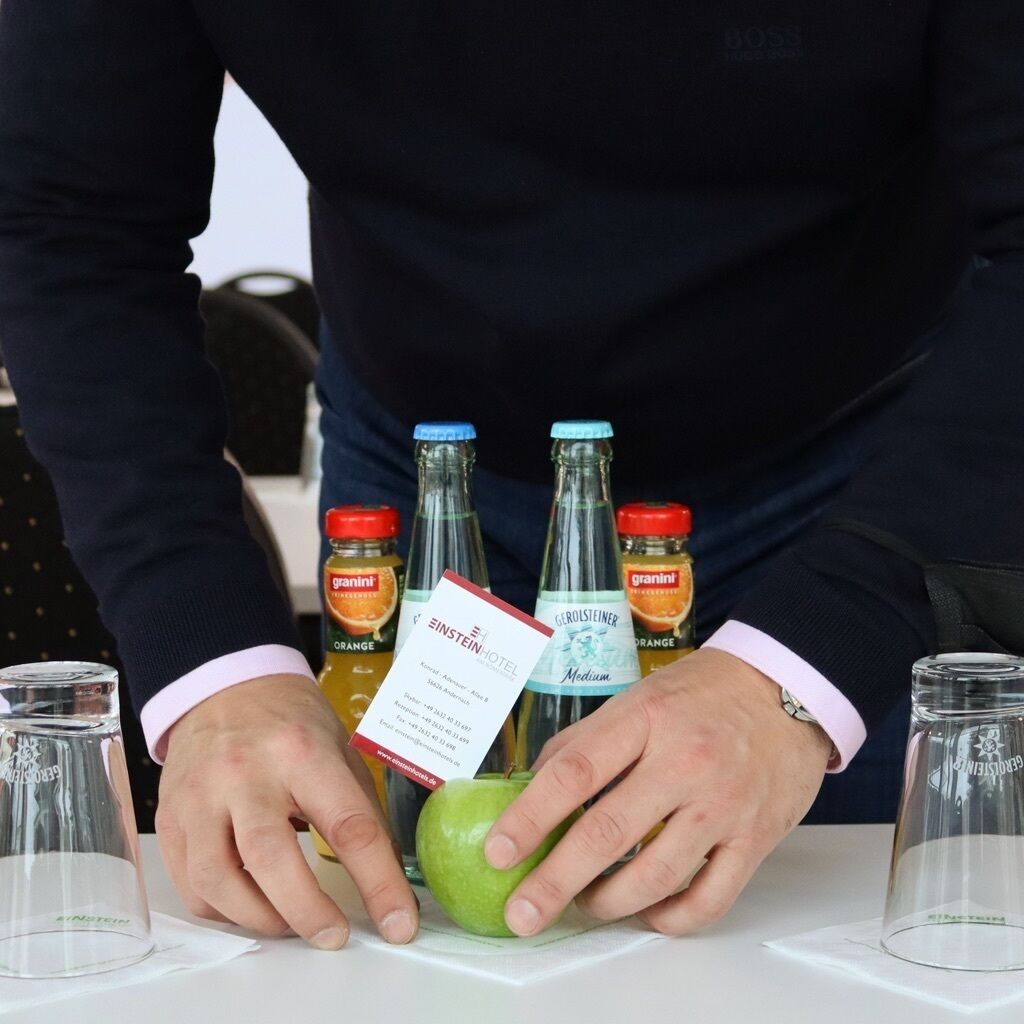 En person holder et grønt æble og to flasker Graninit appelsinjuice, mens en hotelkvittering ligger i forgrunden.