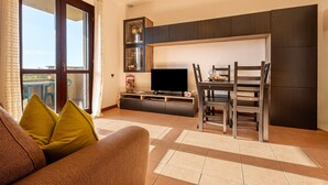 A living room with a sofa, TV, dining table, chairs, and a kitchen area with cabinets and a window. - Terrazza Catabbio – Relax & Saturnia Thermal Baths – Garden and Pool (Catabbio)