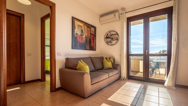 A living room with a sofa, a painting, an air conditioning unit, and a view through a glass door. - Terrazza Catabbio – Relax & Saturnia Thermal Baths – Garden and Pool (Catabbio)