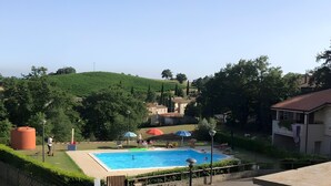 A swimming pool with a view of a green hill, a fenced area, and a building with a red roof. - Terrazza Catabbio – Relax & Saturnia Thermal Baths – Garden and Pool (Catabbio)