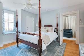 A four-poster bed with a ceiling fan, a window with floral curtains, a blue patterned rug, and a door leading to another room.
