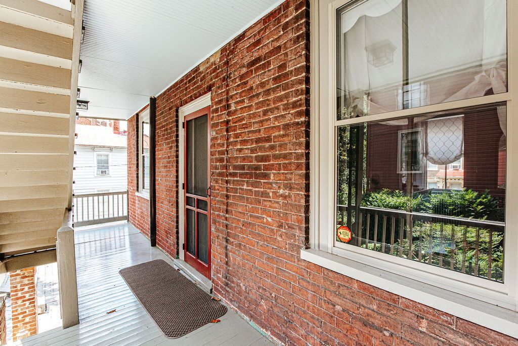 A covered porch with a brick exterior, a red door, and a window with a view of greenery.