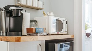 Eine kompakte Küche mit Mikrowelle, Kaffeemaschine und kleinem Kühlschrank. Über der Arbeitsfläche befindet sich ein Regal mit Tassen und einem Einmachglas.