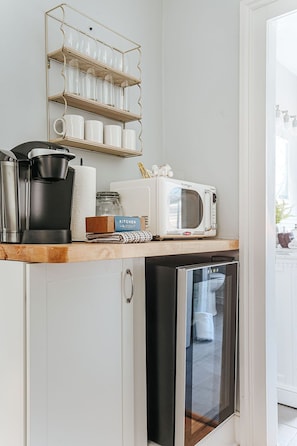 Una cocina compacta con microondas, cafetera y un refrigerador pequeño. Hay una estantería sobre la encimera con tazas y un tarro.