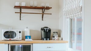Eine kompakte Küche mit Mikrowelle, Kaffeemaschine und kleinem Kühlschrank. Eine Holzhilfe über der Arbeitsfläche beherbergt Tassen und Gläser.