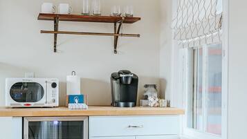 A compact kitchen with a microwave, coffee maker, and a small refrigerator. There is a wooden shelf above the counter holding cups and glasses.