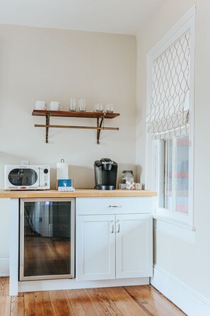 Una cocina compacta con microondas, cafetera y refrigerador pequeño. Hay un estante de madera sobre la encimera con tazas y vasos.