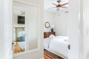 Un dormitorio con cama, ventilador de techo, mesita de noche con lámpara, espejo y televisor en la pared.