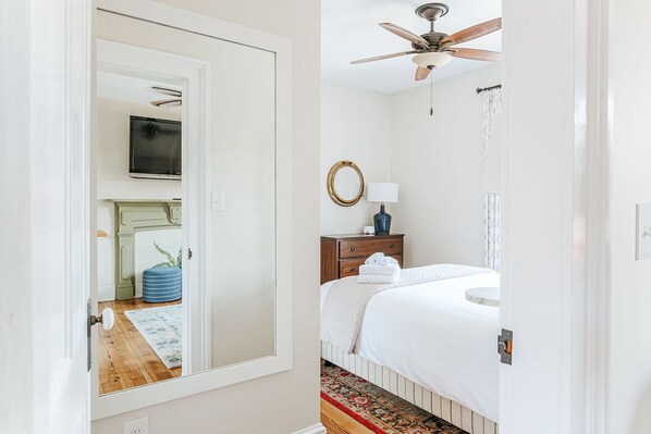 A bedroom with a bed, a ceiling fan, a bedside table with a lamp, a mirror, and a television on the wall.