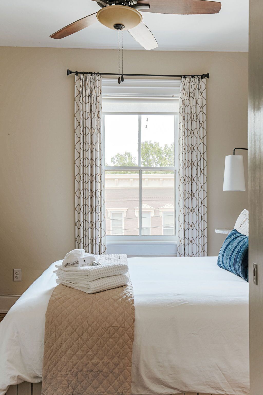A bedroom with a bed, a ceiling fan, a window with curtains, and a lamp.