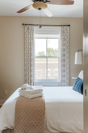 A bedroom with a bed, a ceiling fan, a window with curtains, and a lamp.