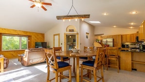 A spacious living room with a large wooden dining table surrounded by chairs, a ceiling fan, and a TV.