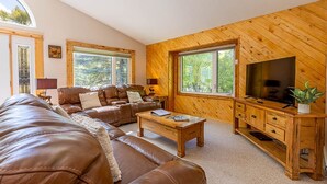 A cozy living room with a brown leather sofa, a wooden coffee table, and a TV on a wooden stand. Large windows offer a view of the outdoors.
