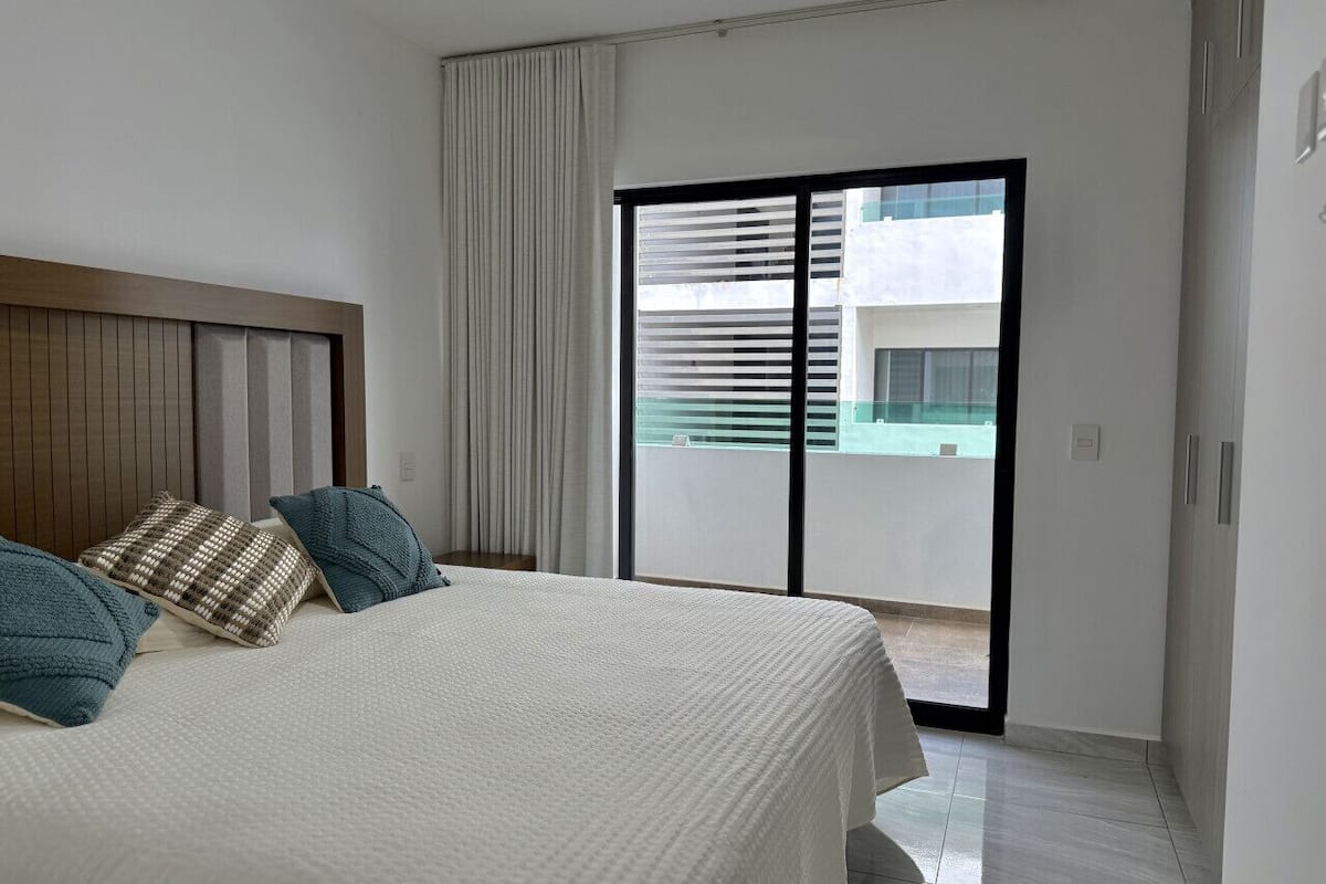 A bedroom with a bed, a headboard, and a view of a building through a window.
