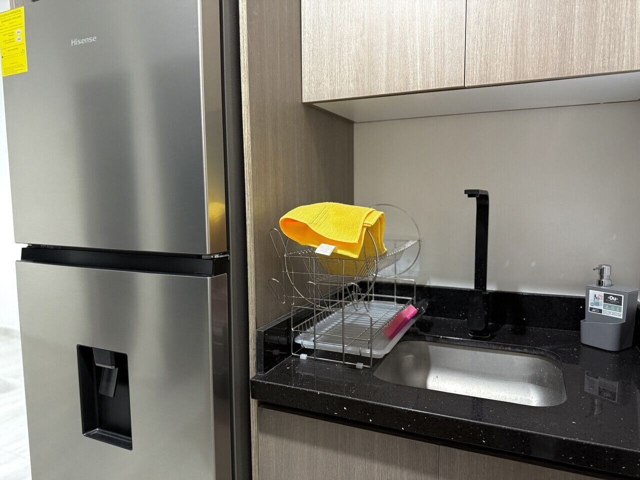 A stainless steel refrigerator with a yellow towel on the door, a dish rack with a yellow towel, a black faucet, and a soap dispenser on a black countertop.