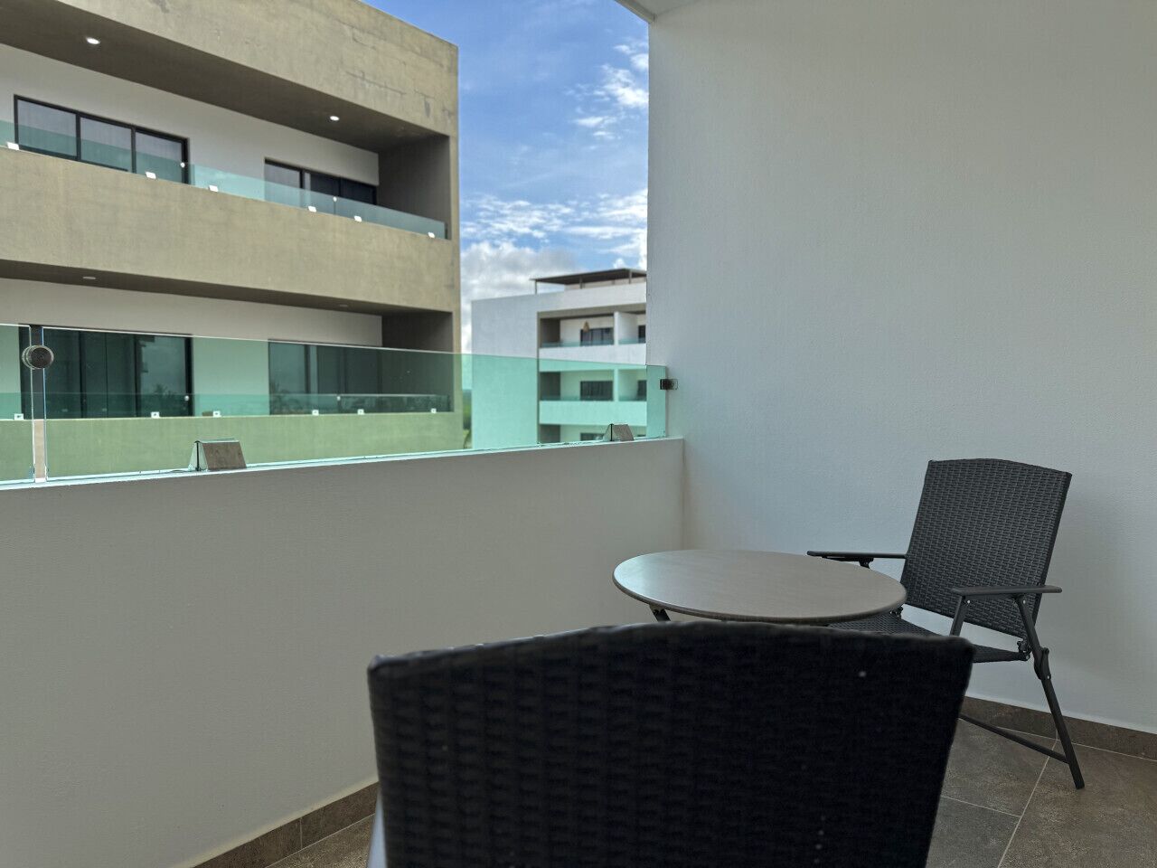 A balcony with a round table and two chairs, overlooking a building with multiple floors and a clear sky.