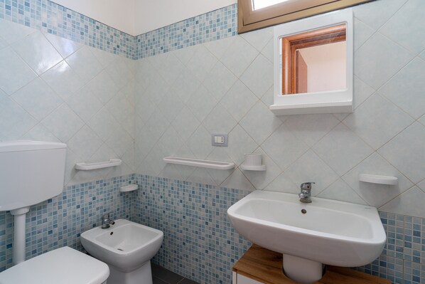 A bathroom with a toilet, bidet, and sink, tiled with blue and white tiles, and a small window above the sink.