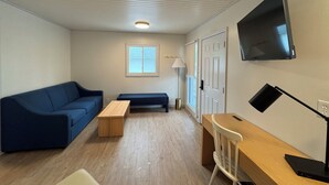A room with a blue sofa, a wooden table, a white chair, a TV mounted on the wall, and a window with blinds. - Waterfall Cottage at GOTL (Geneva)