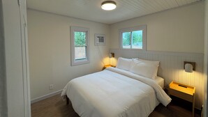 A bedroom with a bed, two windows, a ceiling light, a wall-mounted air conditioner, and a nightstand with a lamp. - Waterfall Cottage at GOTL (Geneva)