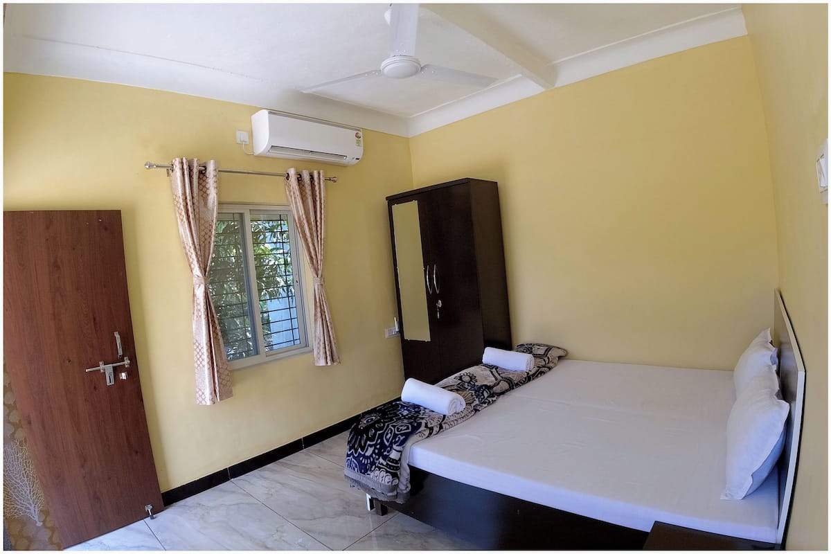 A bedroom with a bed, a wardrobe, a window with curtains, an air conditioning unit, and a ceiling fan.