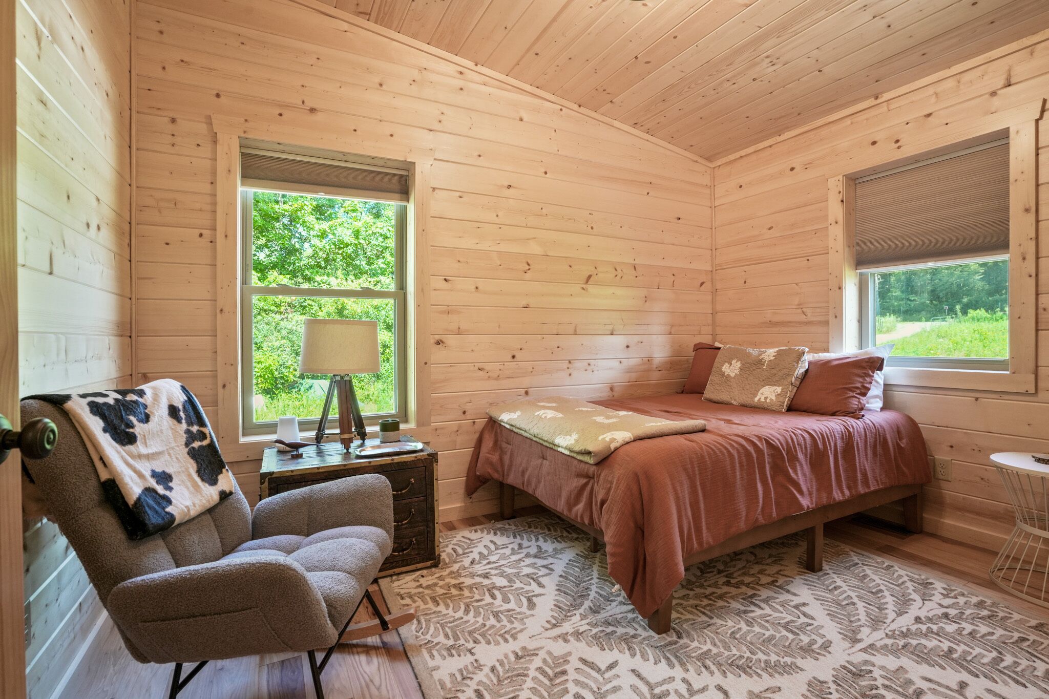 A cozy wooden cabin bedroom with a bed, a chair, a nightstand, and a window with a view of greenery.
