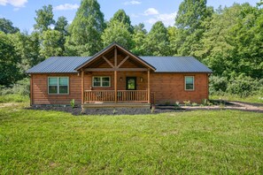 Une cabane en rondins dotée d’un porche, entourée d’un champ verdoyant et d’une forêt dense.