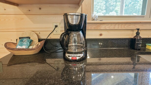 A coffee maker on a kitchen counter with a wooden cabinet above and a window with a view of trees. - Coyote Creek | Hocking Hills, 40 Acre Retreat (McArthur)