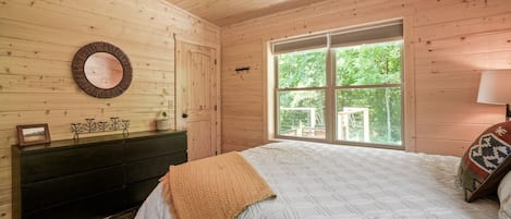 Un dormitorio con techo de madera, un ventanal con vistas a la vegetación, una cama con colcha blanca y un aparador de madera oscura.