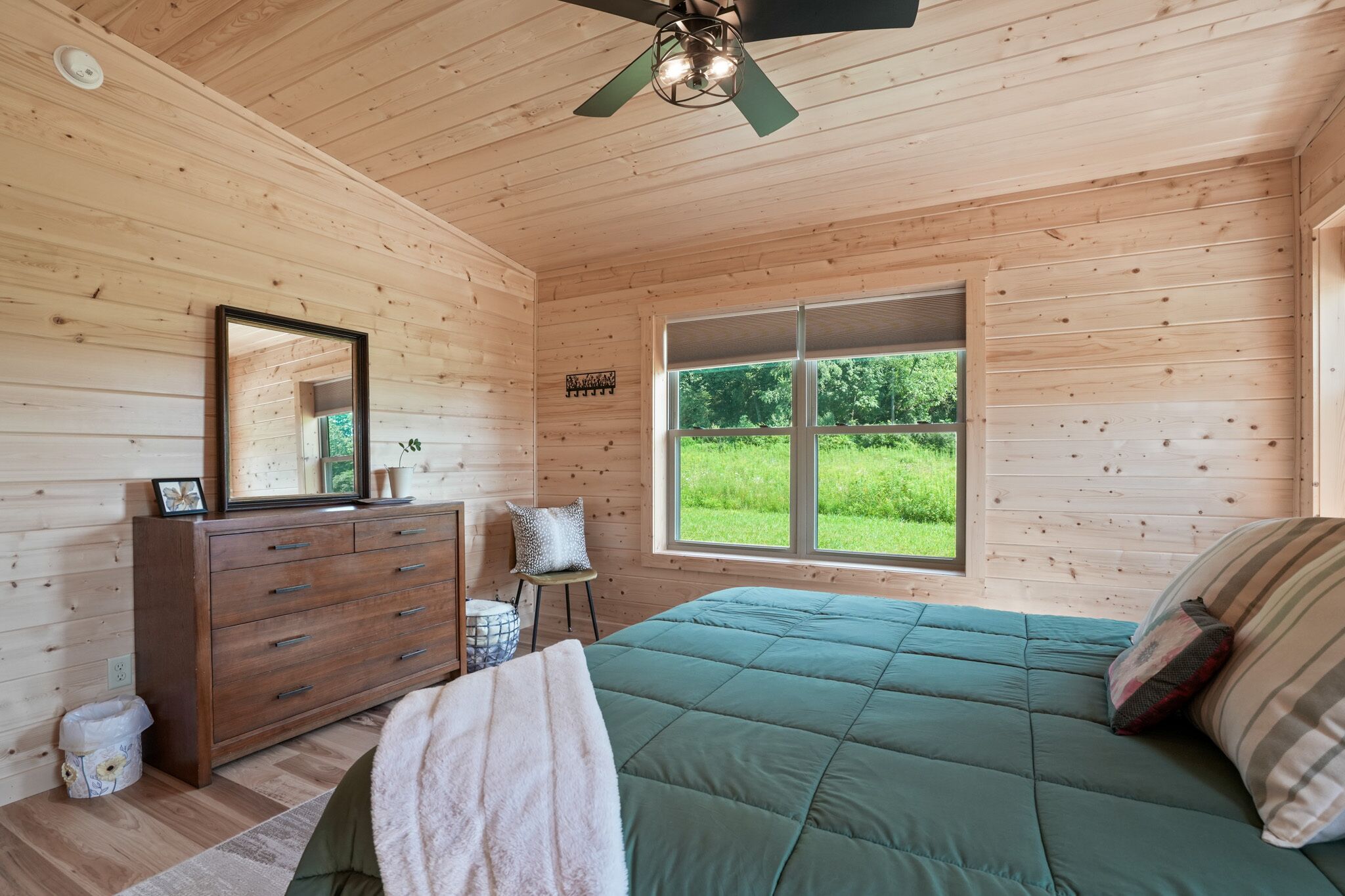 A wooden cabin bedroom with a bed, dresser, mirror, and a window with a view of greenery.