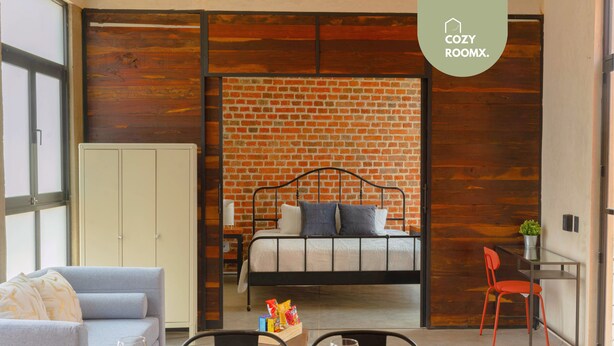 A modern room with a brick wall, a bed, a grey sofa, and a dining area with chairs and a table. - Style & Comfort  A/C Loft + Terrace + Pets (Oaxaca de Juárez)