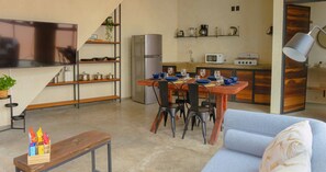 A modern living room with a sofa, a wooden coffee table, and a TV. There is a kitchen area with a refrigerator, microwave, and a dining table set for four.
