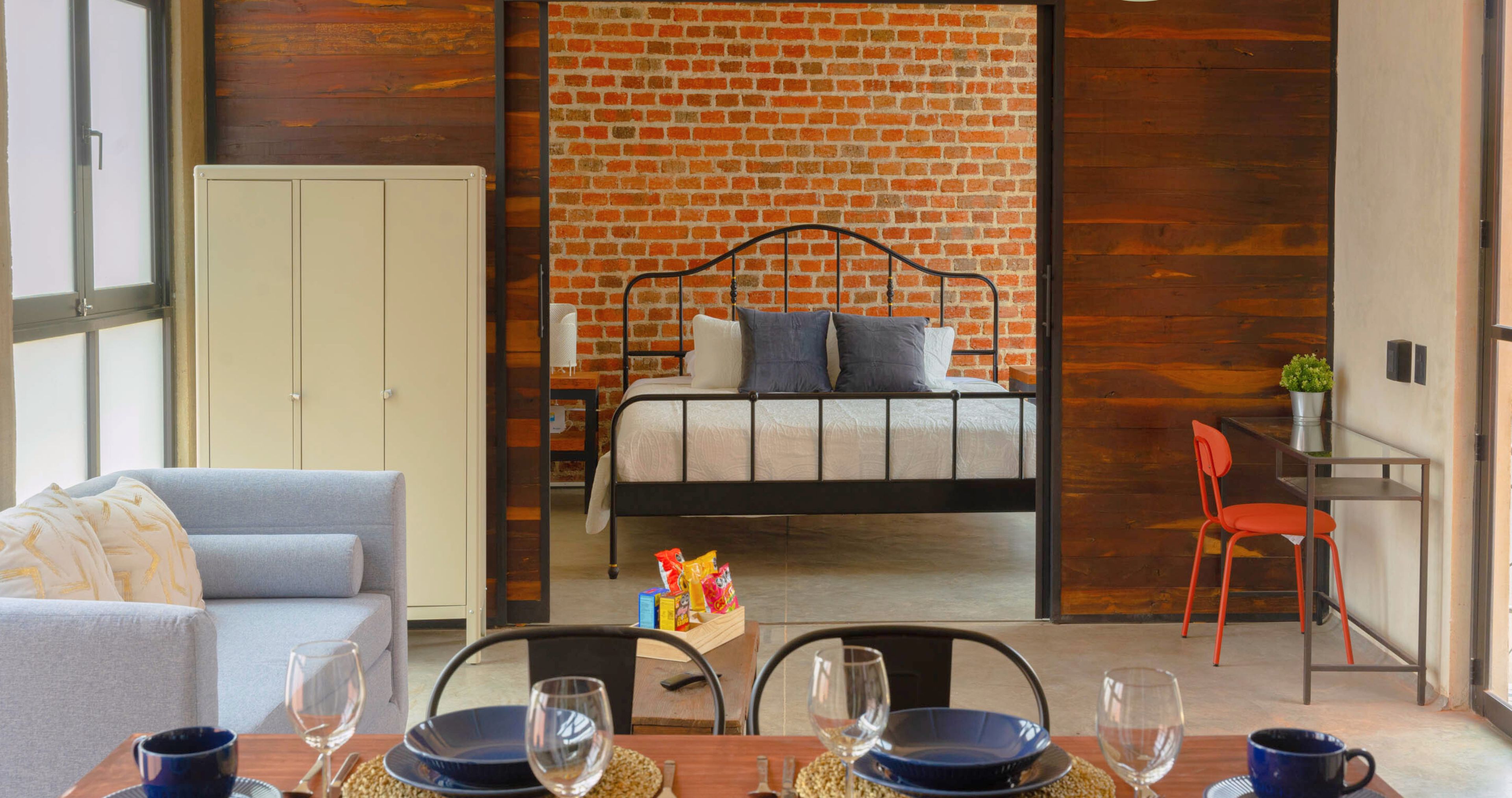 A modern room with a brick wall, a bed, a grey sofa, and a dining area with chairs and a table.