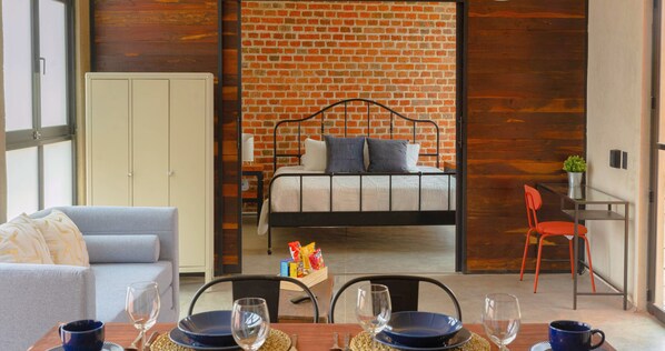 A modern room with a brick wall, a bed, a grey sofa, and a dining area with chairs and a table.