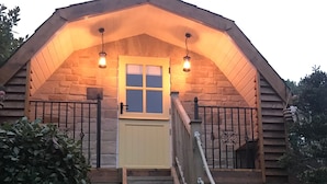 Una cabaña de troncos con una escalera de madera que conduce a una puerta, un pequeño porche con barandilla y dos faroles colgantes.